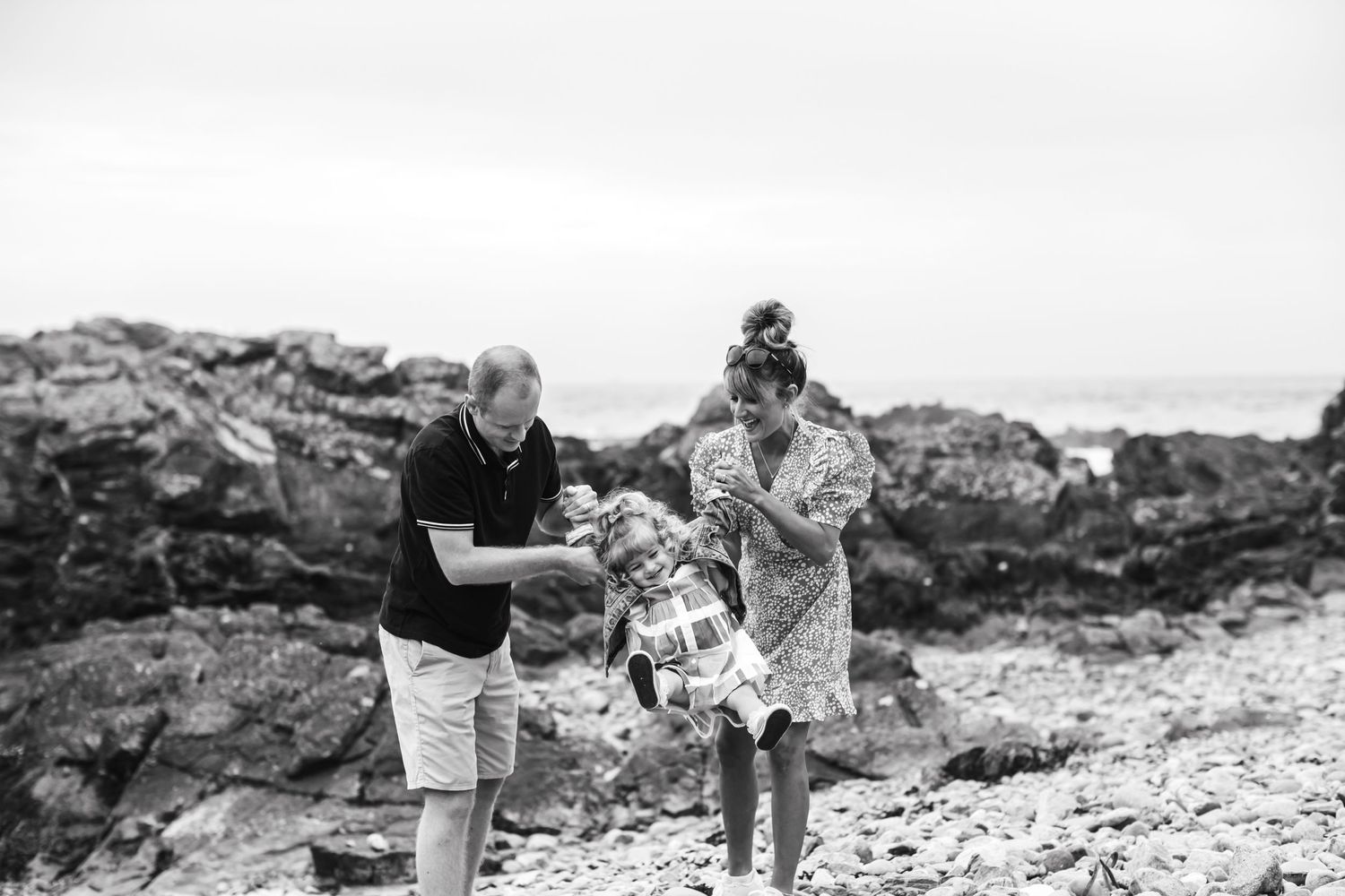 Black and white photo of a family playing on a rocky beach, parents swinging their young child between them.