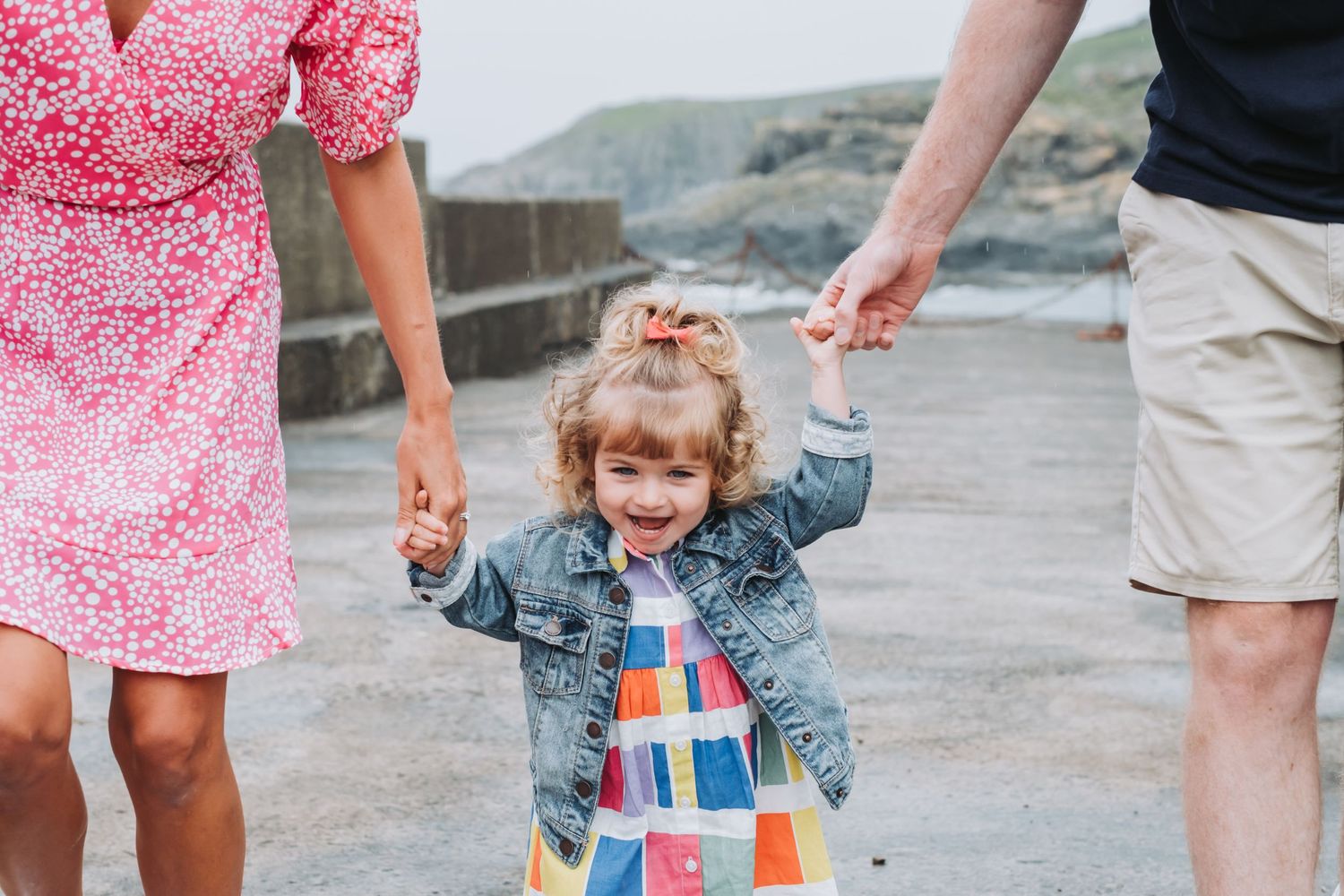 Joyful toddler girl in colorful dress holding parents hands on a seaside promenade.