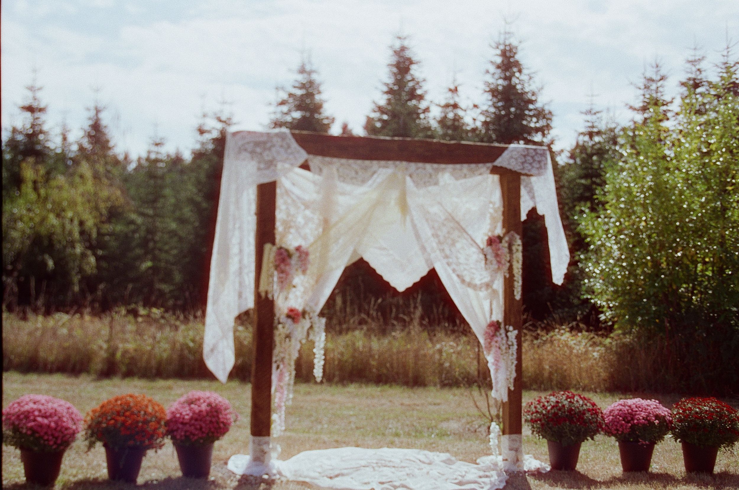 A rustic outdoor wedding ceremony setup with white fabric draped arch and pink floral arrangements in a natural setting.