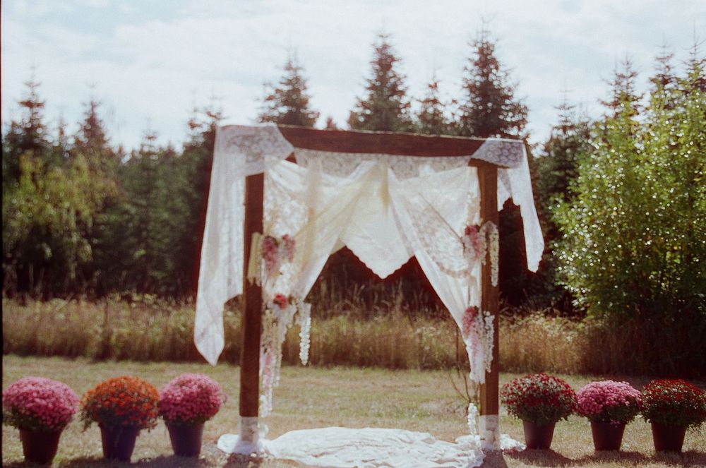 A rustic outdoor wedding ceremony setup with white fabric draped arch and pink floral arrangements in a natural setting.