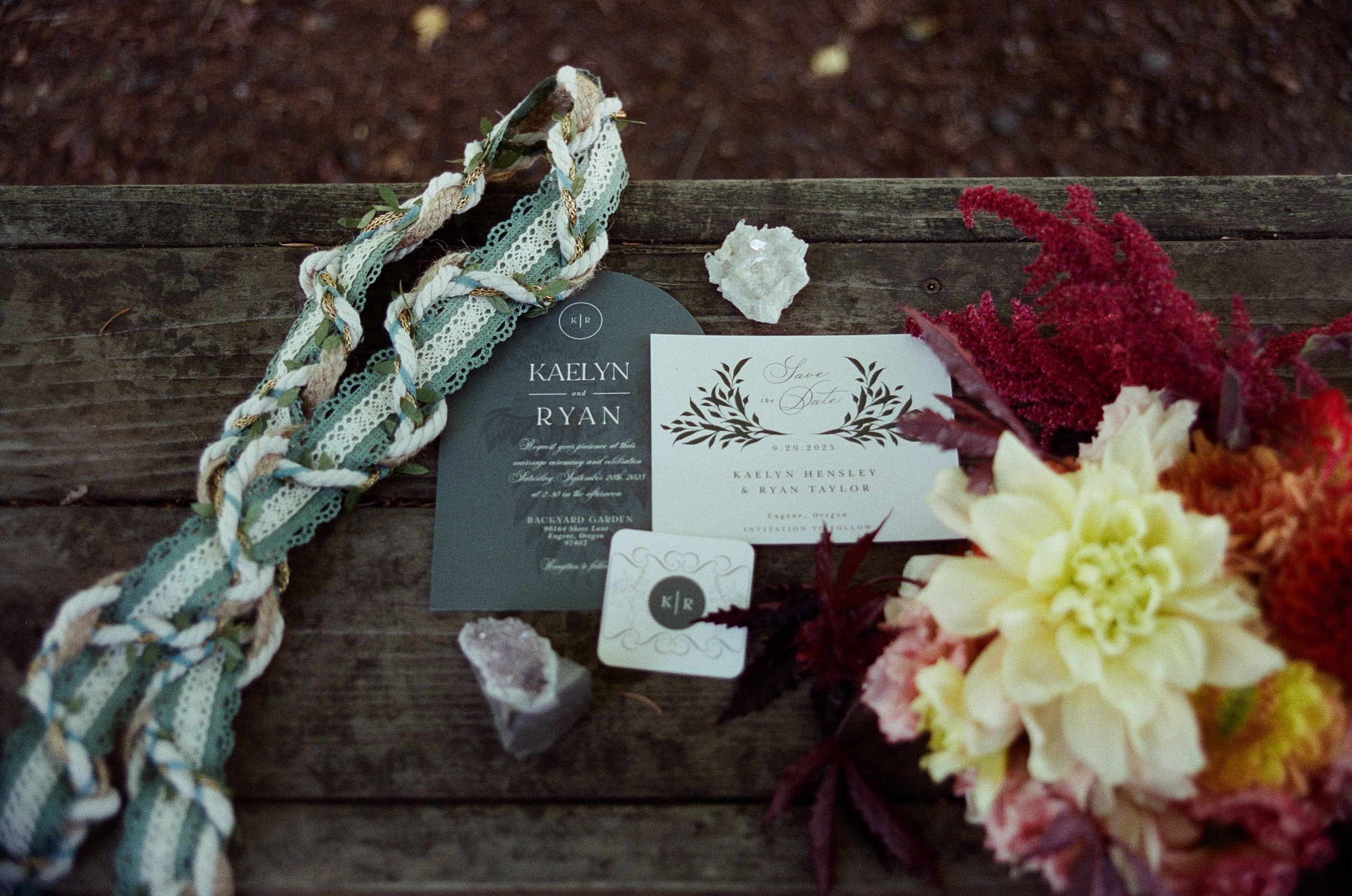 Wedding invitation with floral design displayed on rustic wood alongside crystals and colorful flowers.