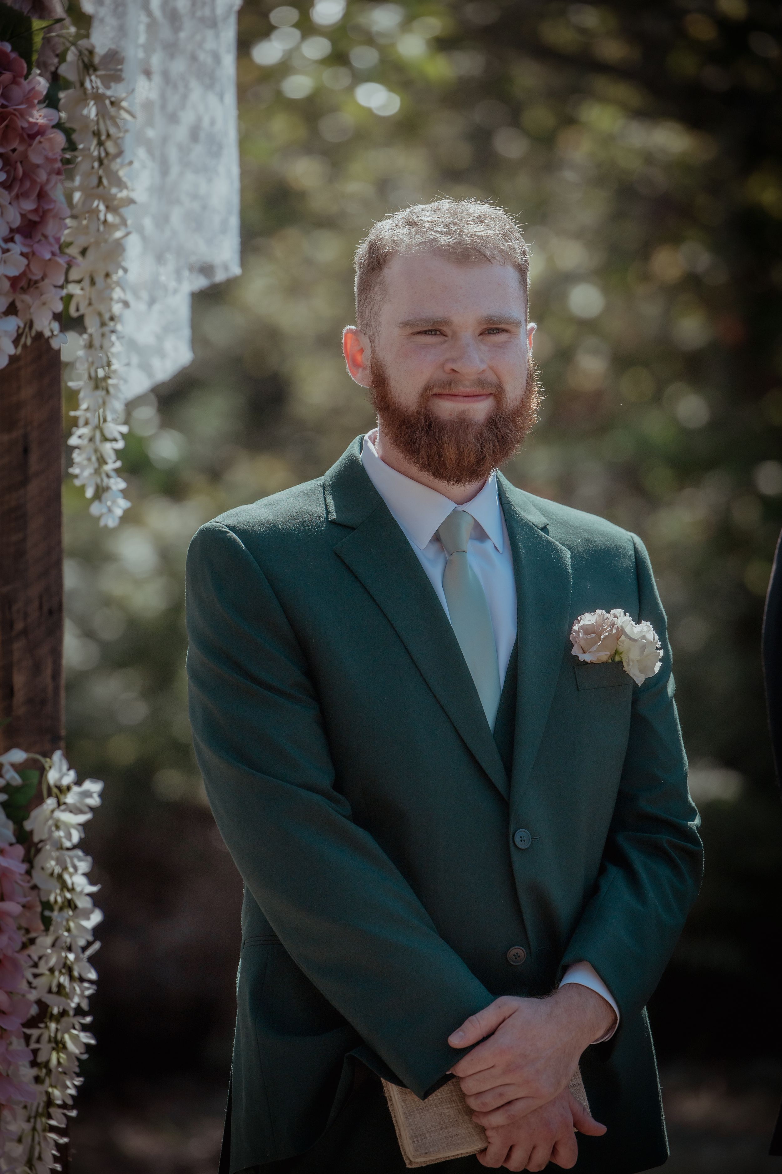Groom in a dark green suit with a red beard poses during an outdoor wedding ceremony.