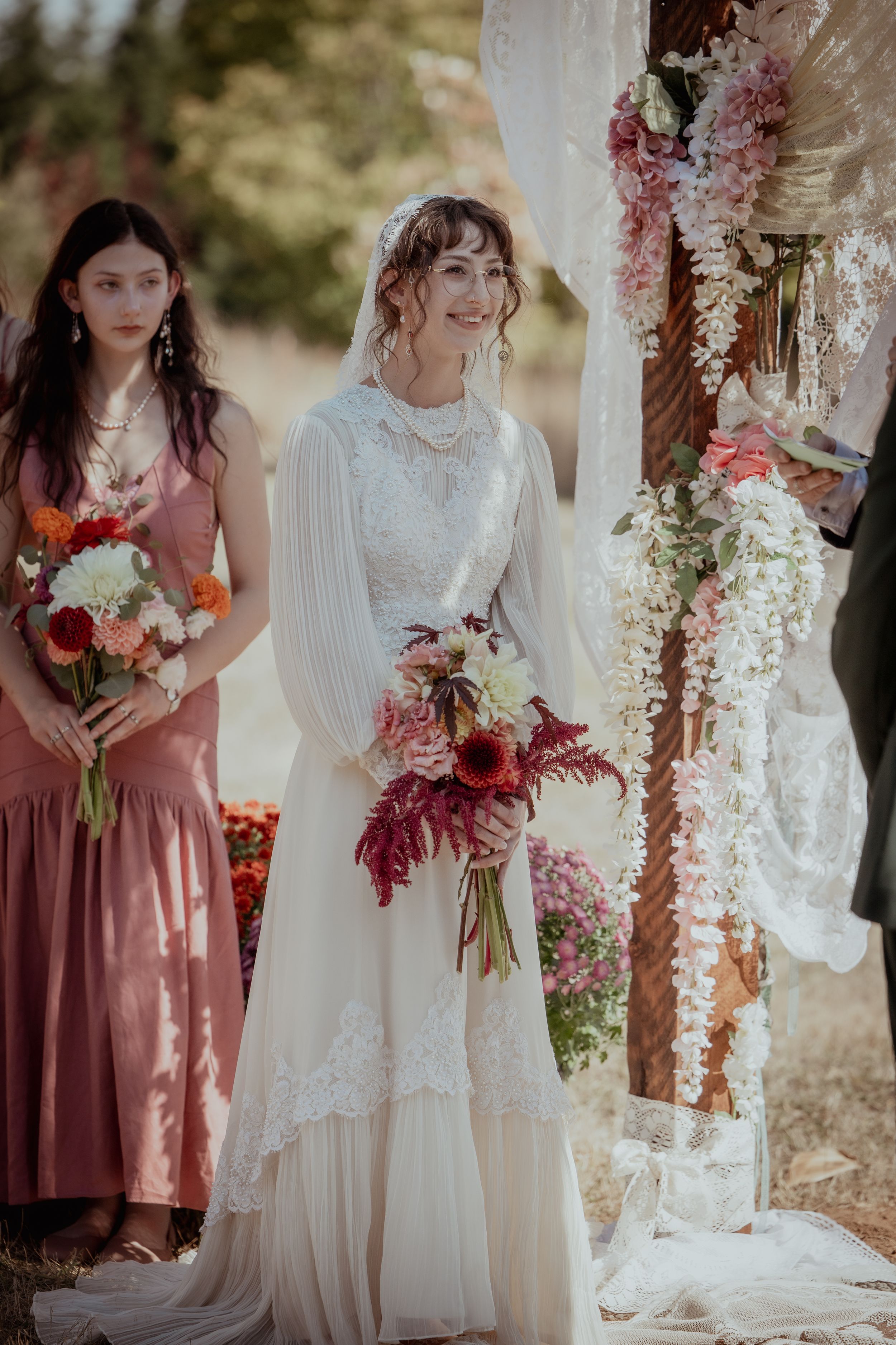 Bridesmaids in burgundy dresses stand with others in white gowns holding vibrant floral bouquets at an outdoor wedding.