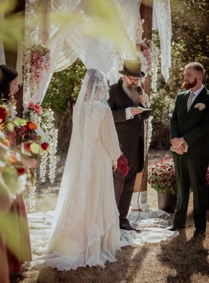 Wedding ceremony under trees with floral decorations and white fabric draping creating a romantic outdoor setting.