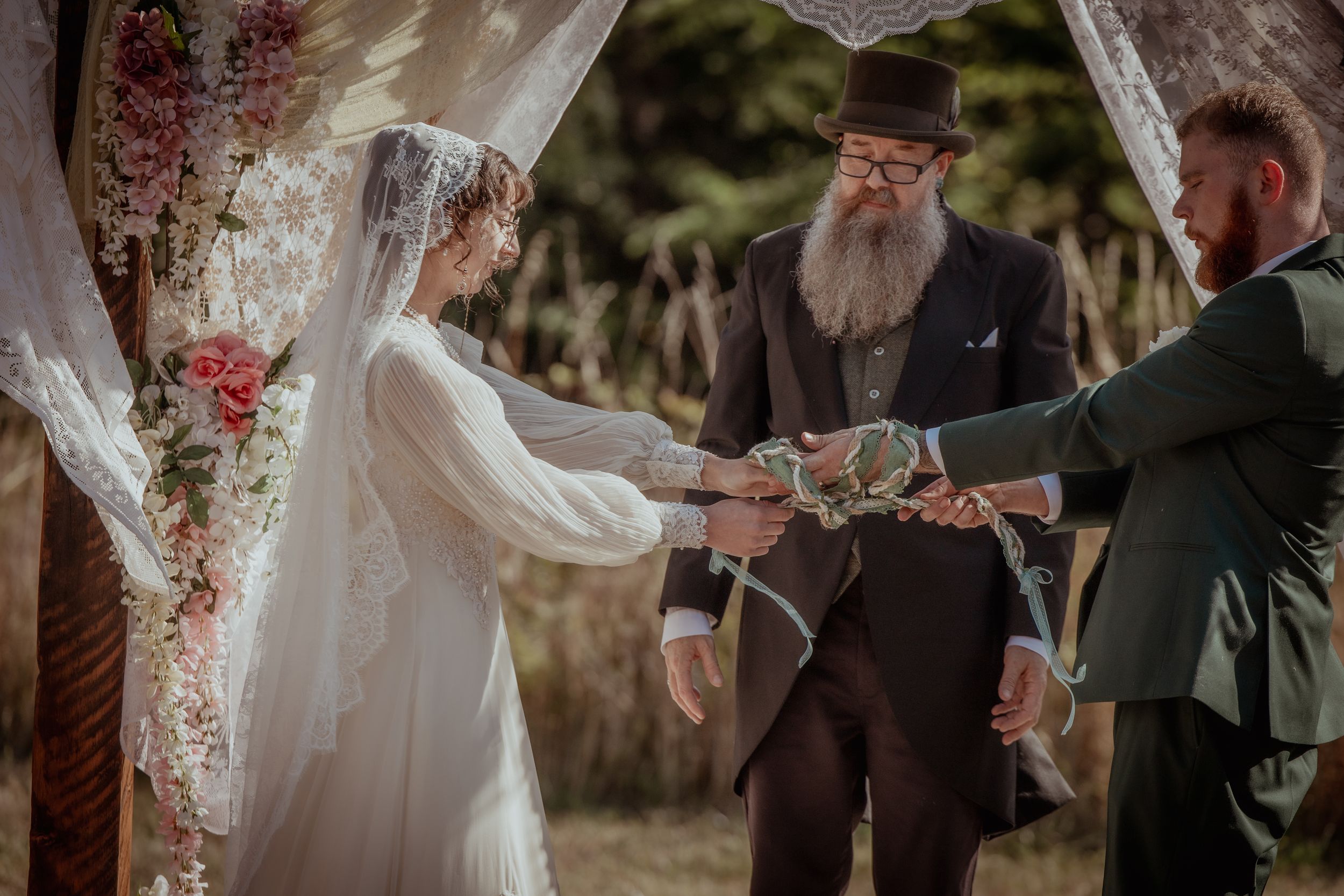 Wedding ceremony guests seated outdoors under floral decorations on a sunny day.
