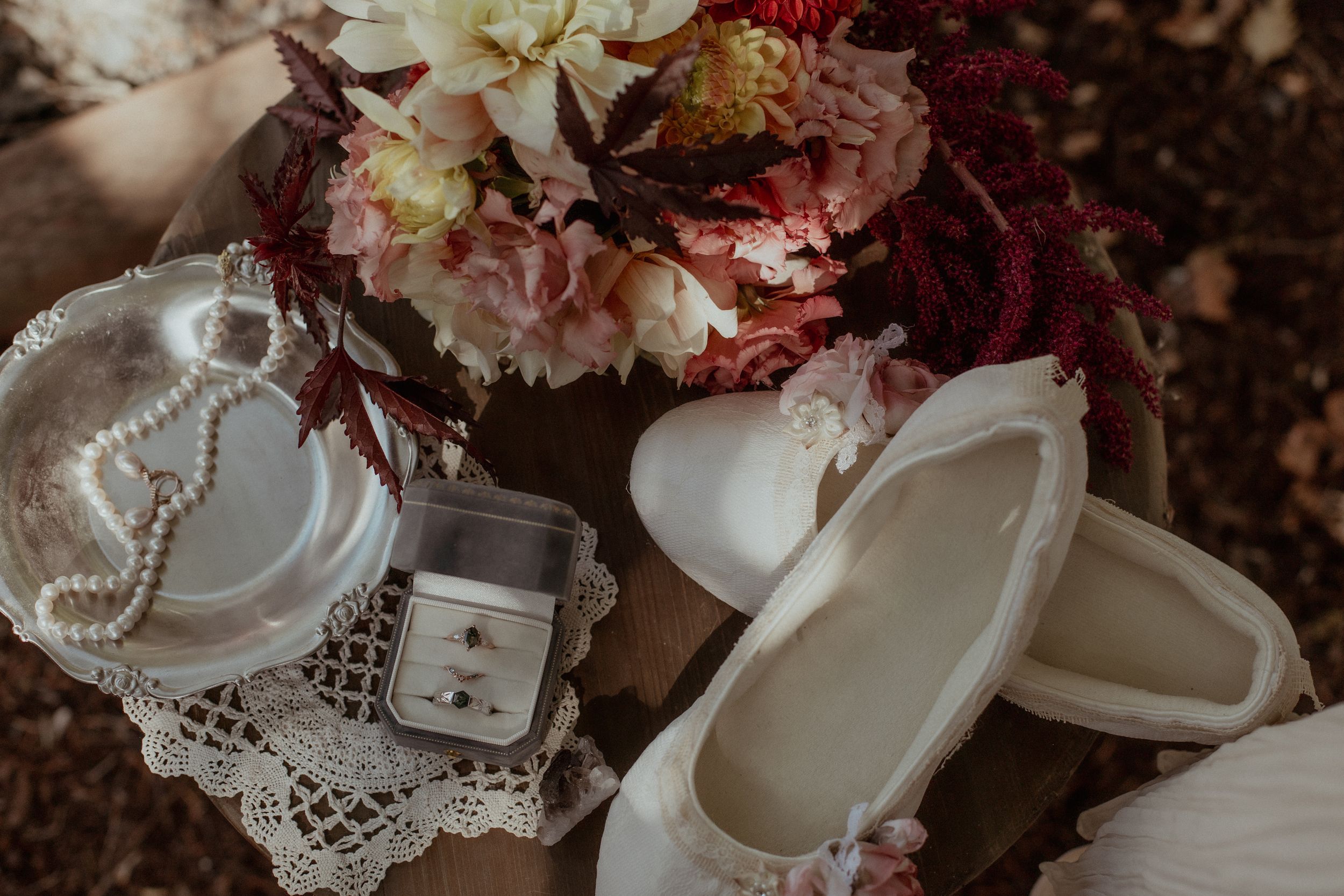 Wedding details including white shoes, lace trim, burgundy flowers, and engagement ring arranged artistically on dark surface.