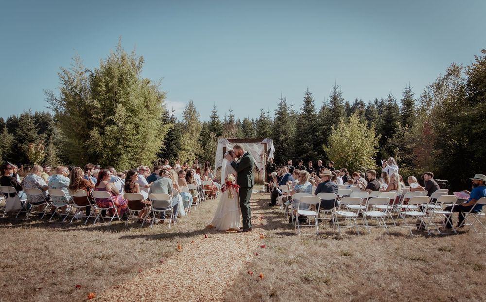 A panoramic view of a sunny outdoor event with people gathered on chairs in a rustic forest setting.