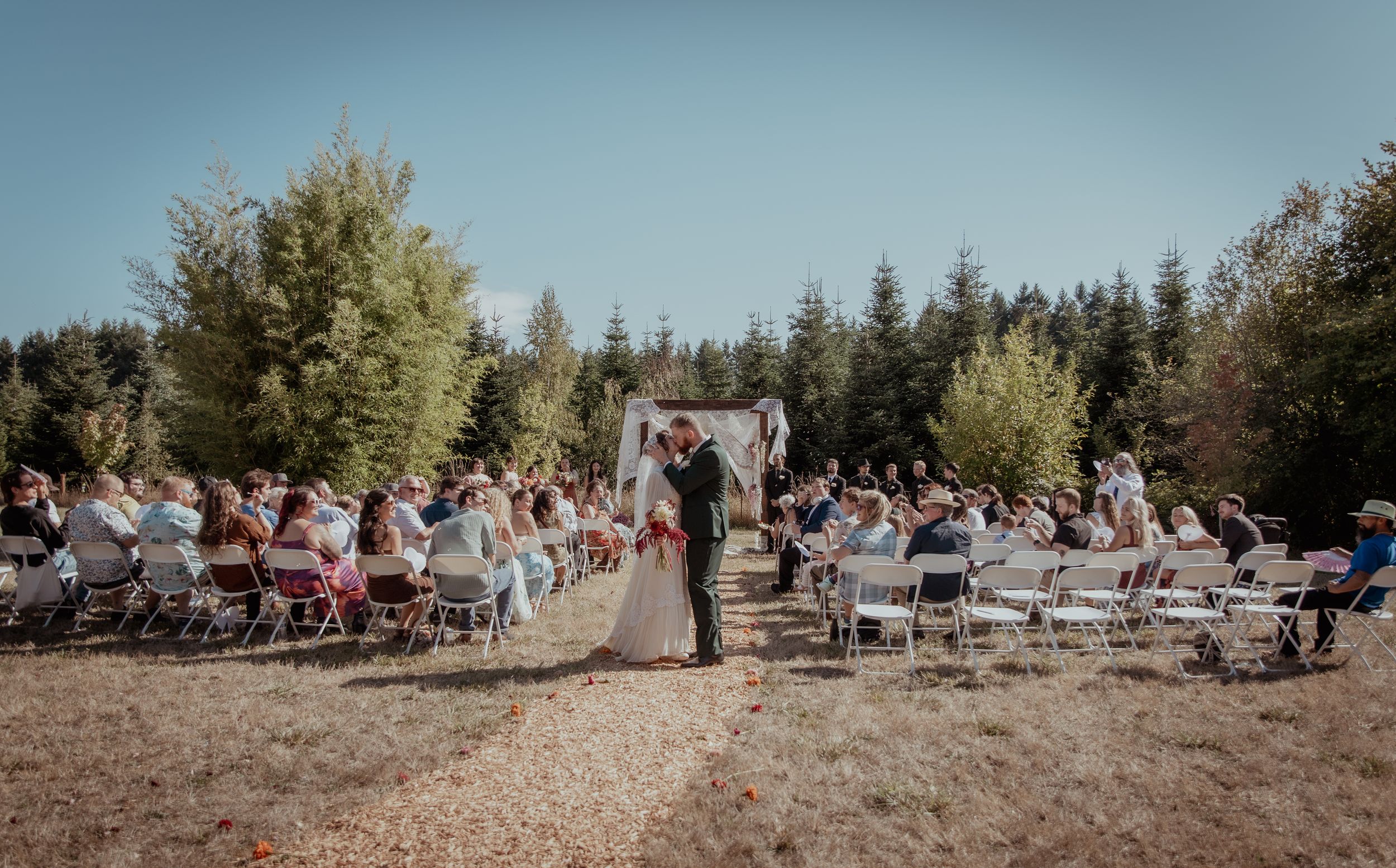 A panoramic view of a sunny outdoor event with people gathered on chairs in a rustic forest setting.