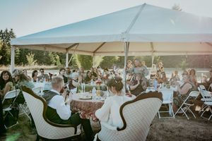 Guests gather under a white tent canopy at an outdoor event with folding chairs and wicker furniture at sunset.