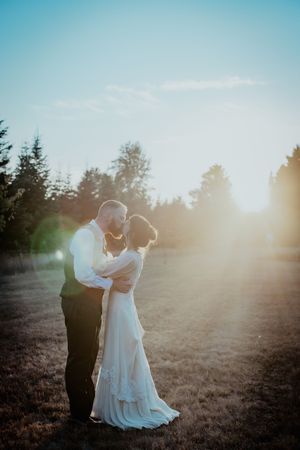 Newlyweds share an intimate moment at sunset in an open field with evergreen trees in the background.