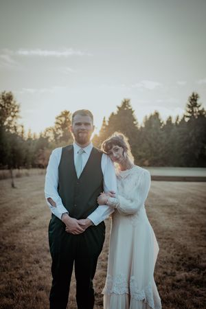 Couple stands together in formal attire during golden hour in an open field with evergreen trees in background.