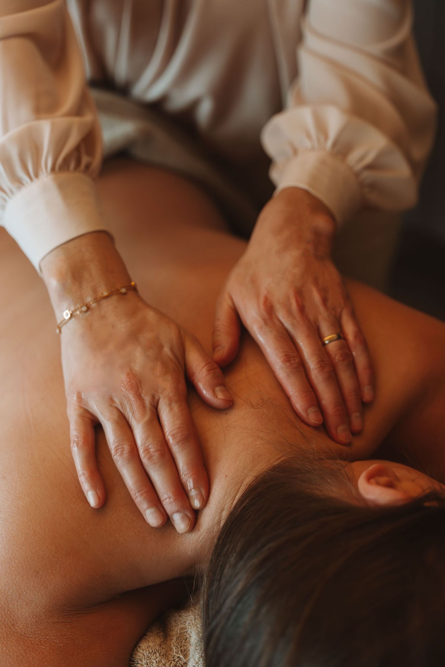 A massage therapist performs therapeutic back massage with hands wearing gold jewelry and white sleeves.