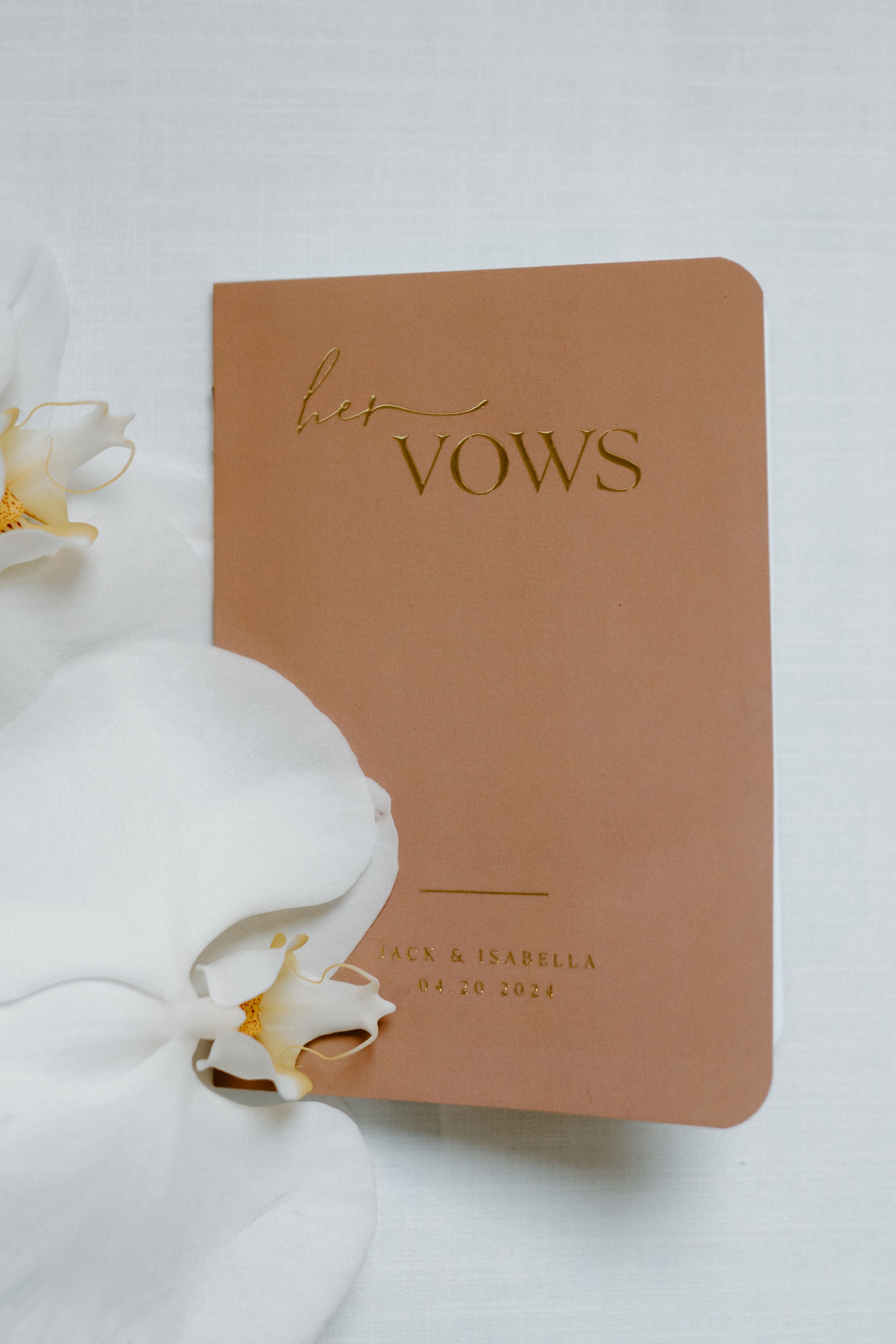 A tan leather wedding vows book with gold text lies beside white orchid flowers on a light background.