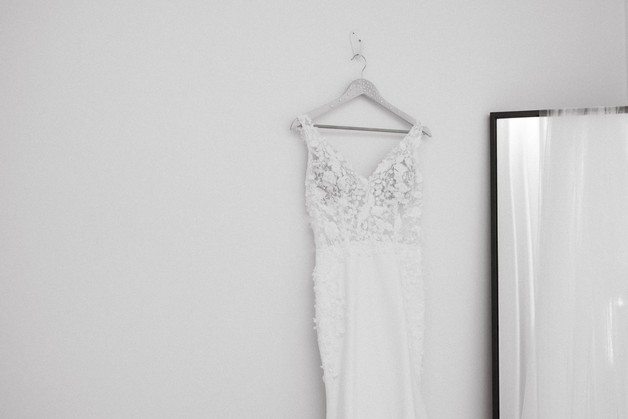 A white lace wedding dress with delicate floral detailing hangs on a hanger against a light gray wall.