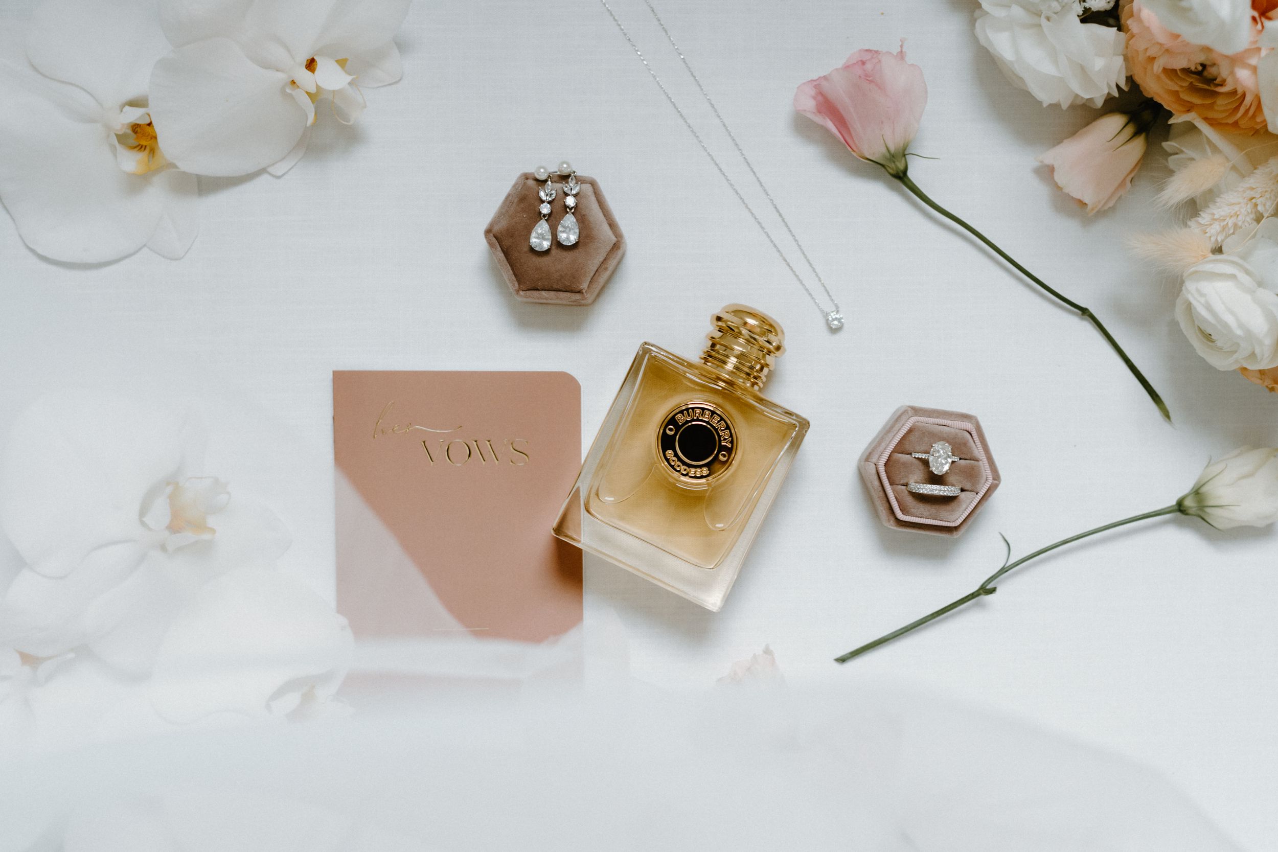 Luxury perfume bottles and cosmetic products arranged with delicate white orchids and pink flowers on a white surface.