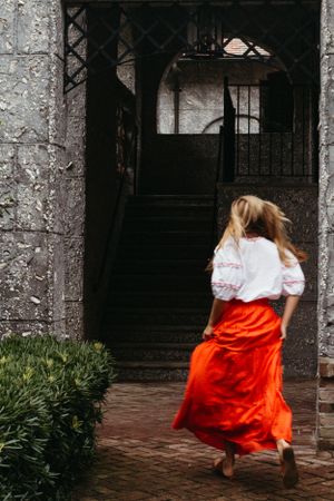 A person in a white blouse and red skirt walks up stone steps into a shadowy archway.