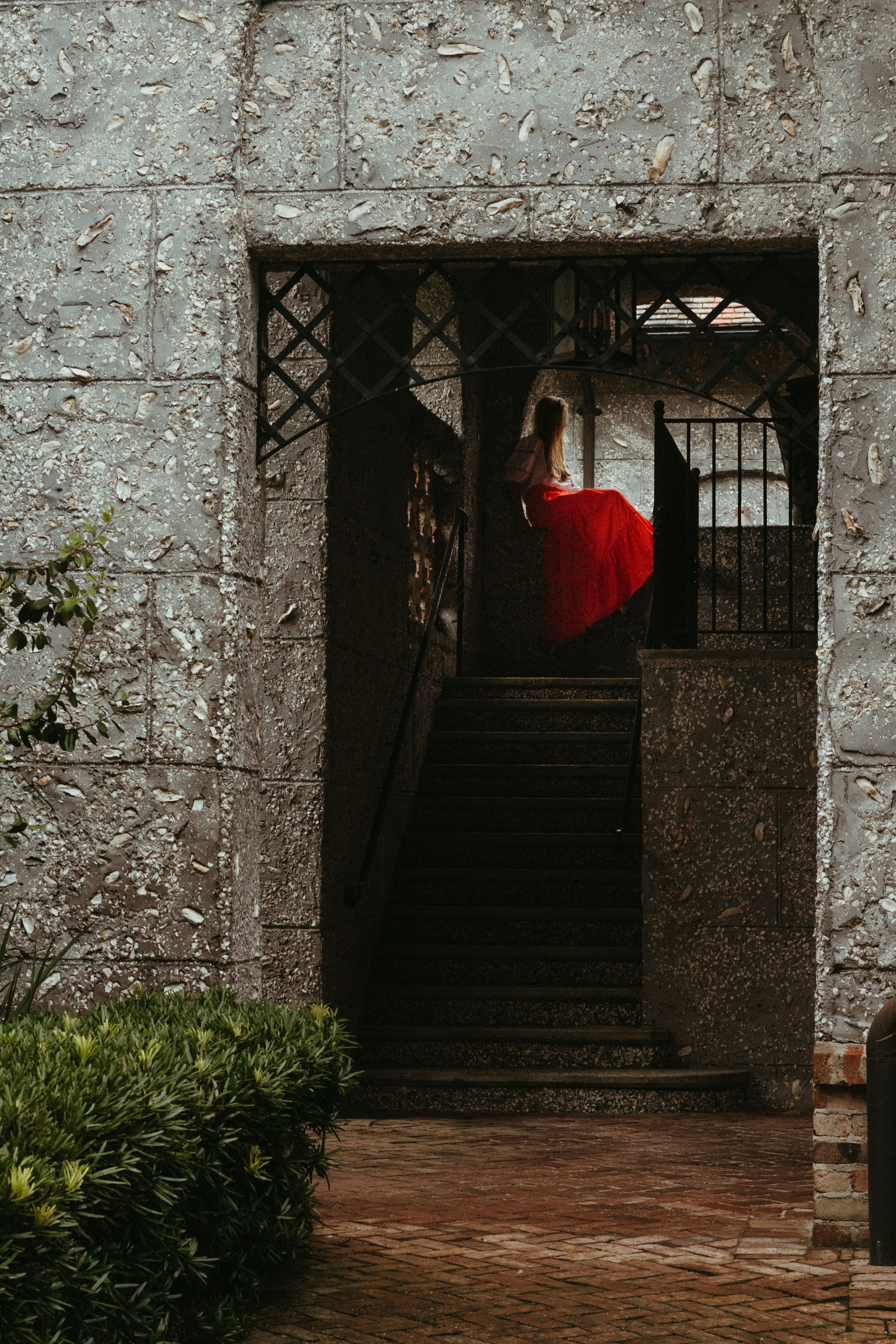 A red dress disappears up dark stone stairs through an ancient doorway with iron gates in a weathered wall.