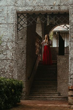 A figure in a flowing red dress stands on stone stairs within a medieval castle archway at dusk.