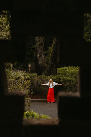 A figure in a red skirt and white top stands with arms outstretched on a dark garden path framed by foliage.