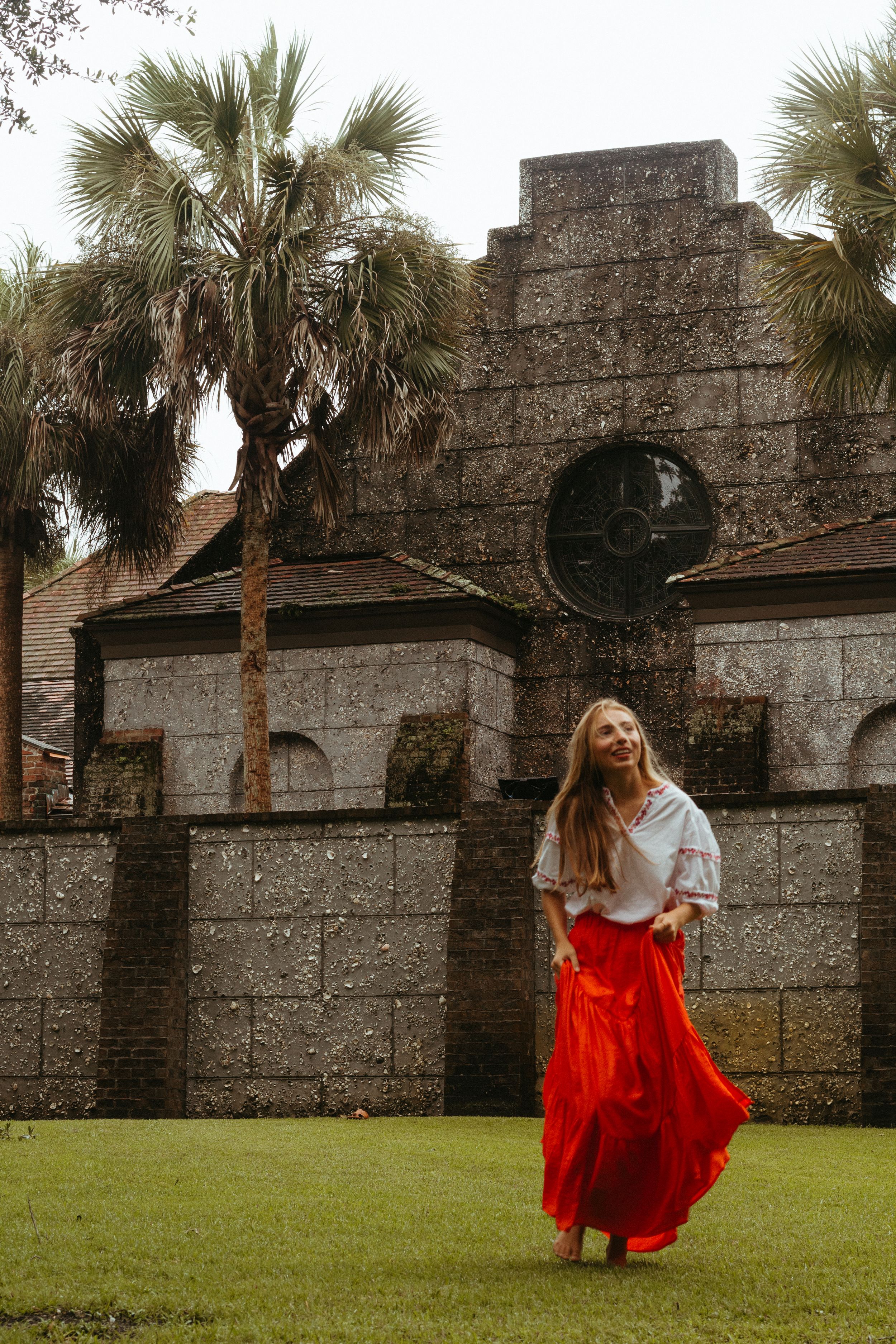 A person in a flowing red skirt and white top stands on grass in front of a stone building with palm trees.