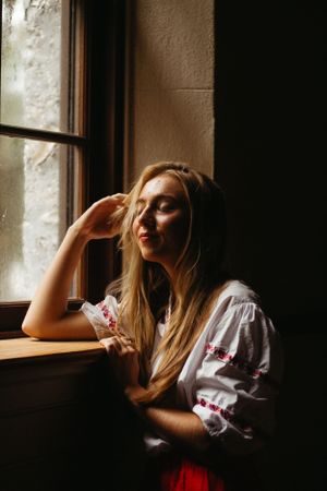 A person in embroidered white peasant blouse sits pensively by window in moody lighting with dramatic shadows.