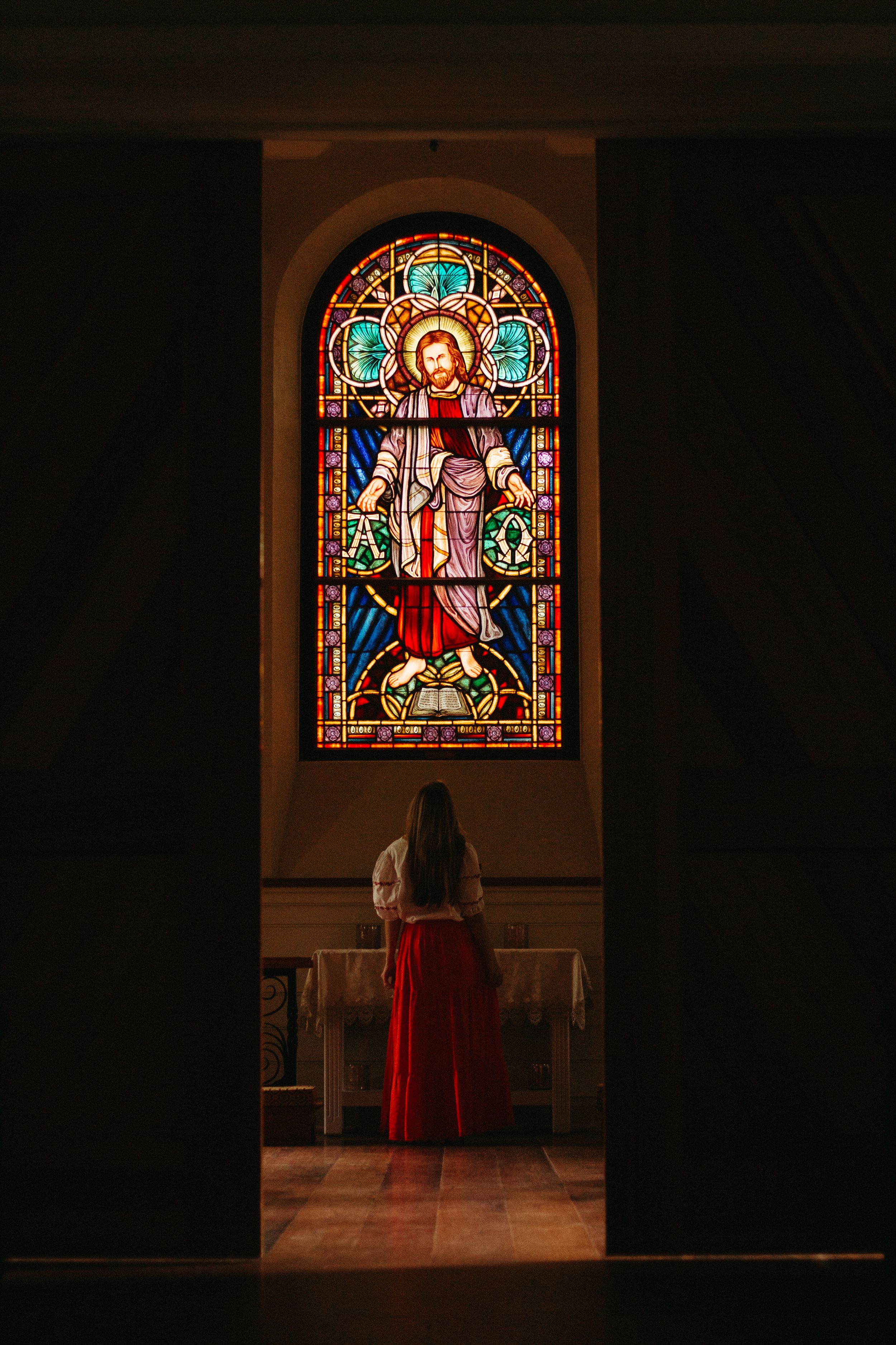 A silhouette gazes up at a colorful stained glass window in a dark church interior.