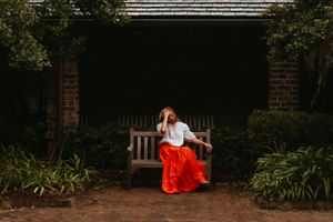 A figure in a flowing red skirt sits on a wooden bench surrounded by greenery.