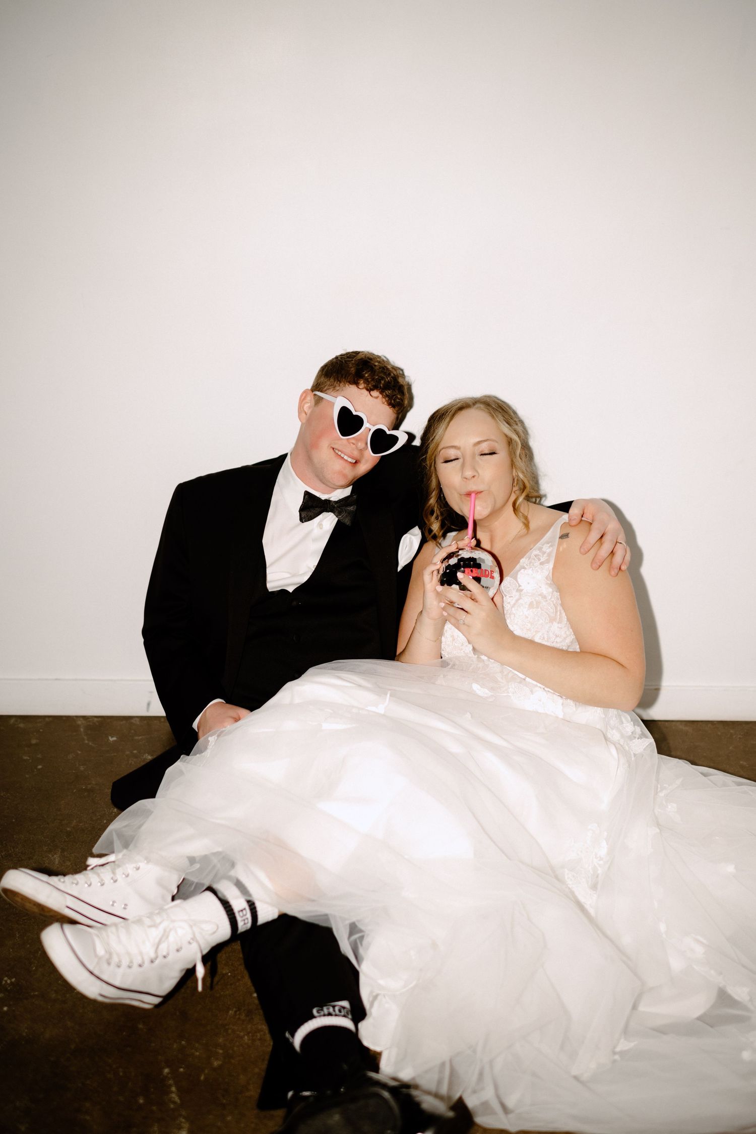 Fun wedding portrait of couple in sunglasses sitting casually against white wall.