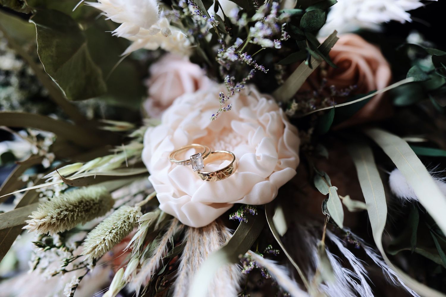 Wedding rings displayed on a light pink peony bloom surrounded by greenery and dried flowers in a romantic arrangement.