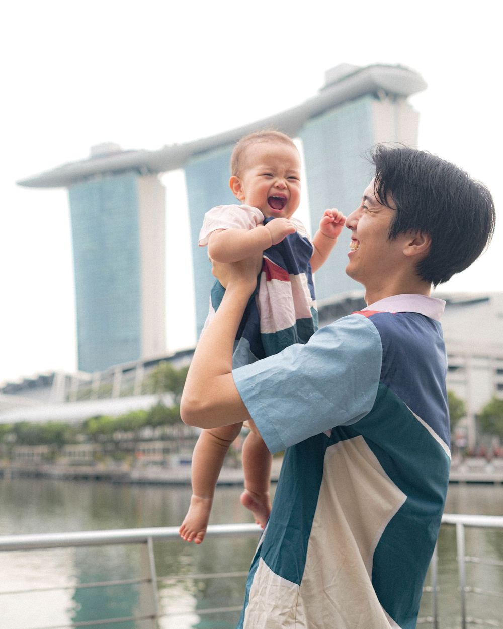 A parent lifts a cheerful baby high with Singapore's Marina Bay Sands in view.