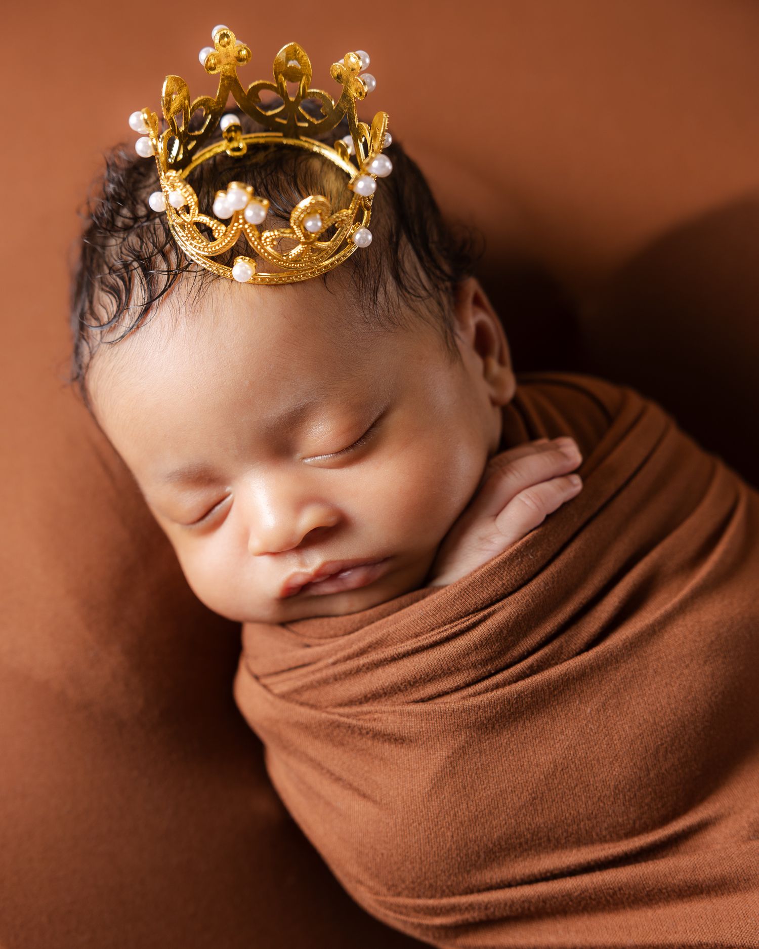 Sleeping baby wrapped in brown fabric wearing a decorative gold crown on brown background.