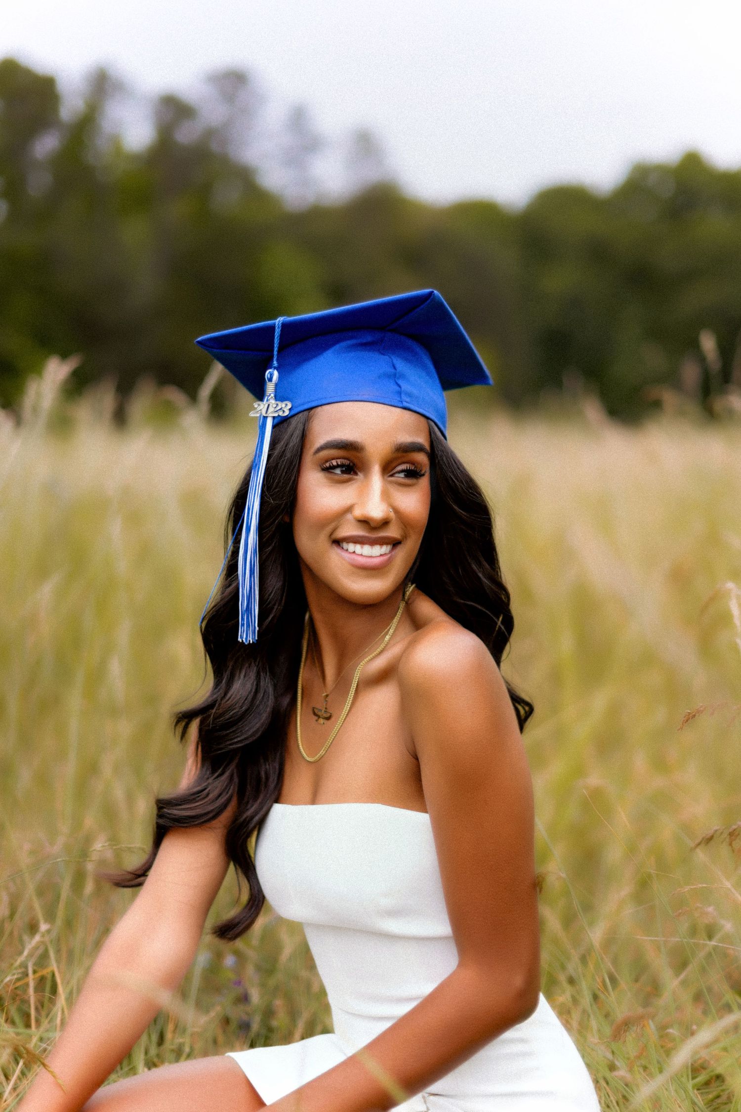 A graduate wearing a blue cap and white dress sits in a sunlit field of tall grass.