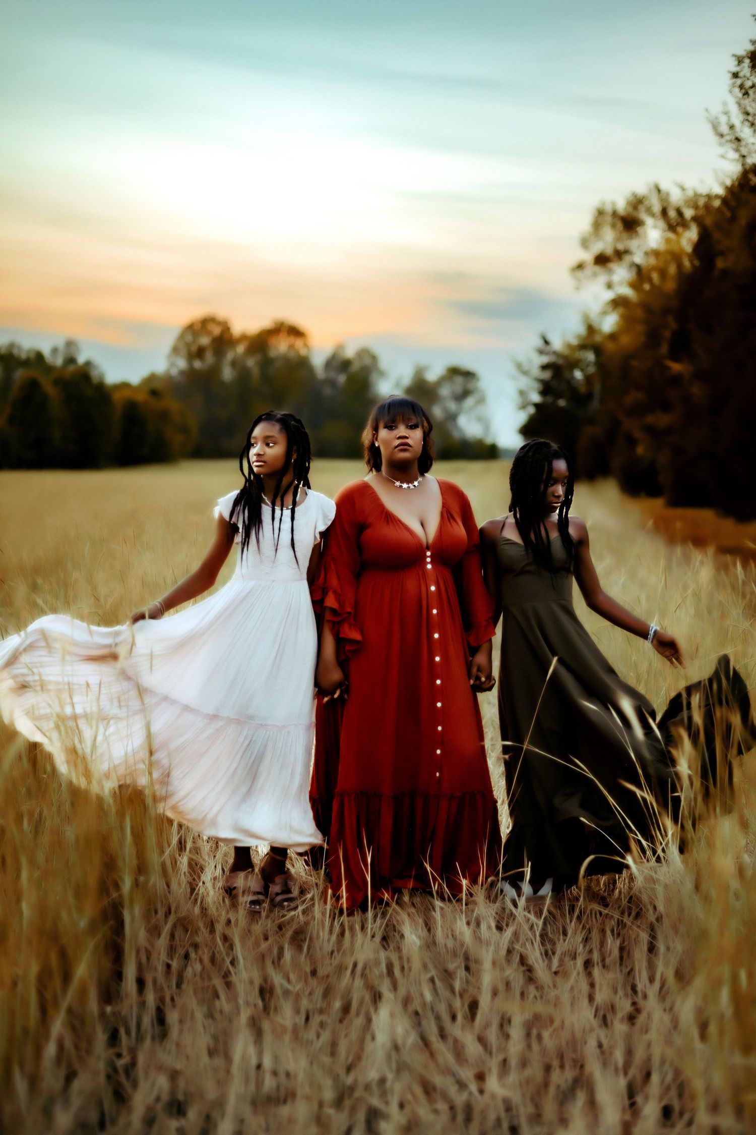 Three women in flowing dresses stand together in a golden wheat field at sunset, creating an ethereal autumn scene.