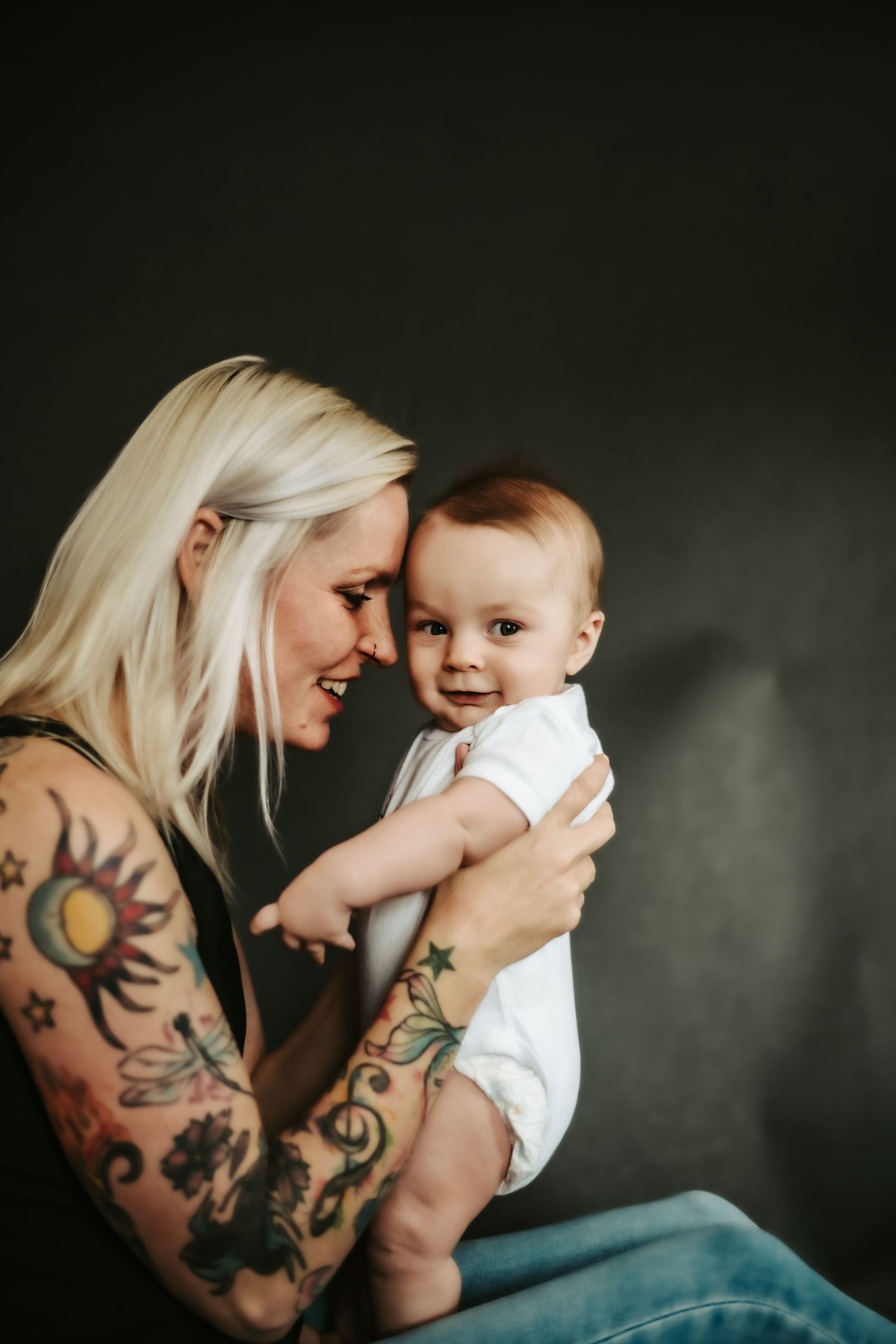 A woman with platinum blonde hair and colorful tattoos holds a smiling baby in white onesie against dark backdrop.