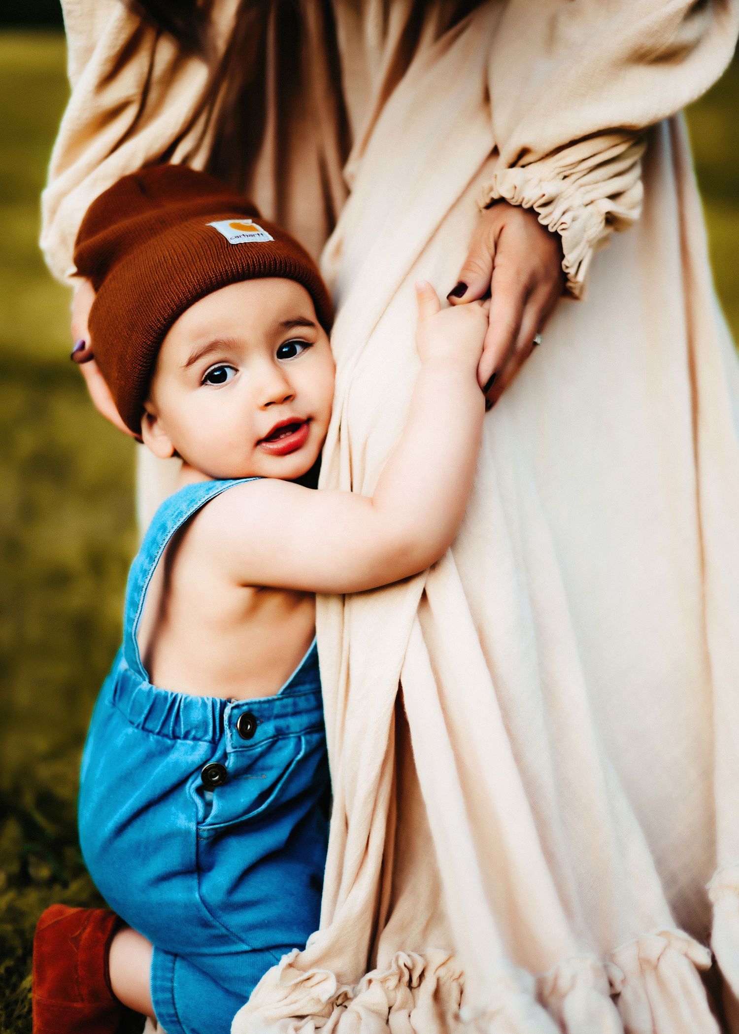 A joyful baby wearing a brown beanie and blue overalls holds onto a beige flowing dress outdoors.