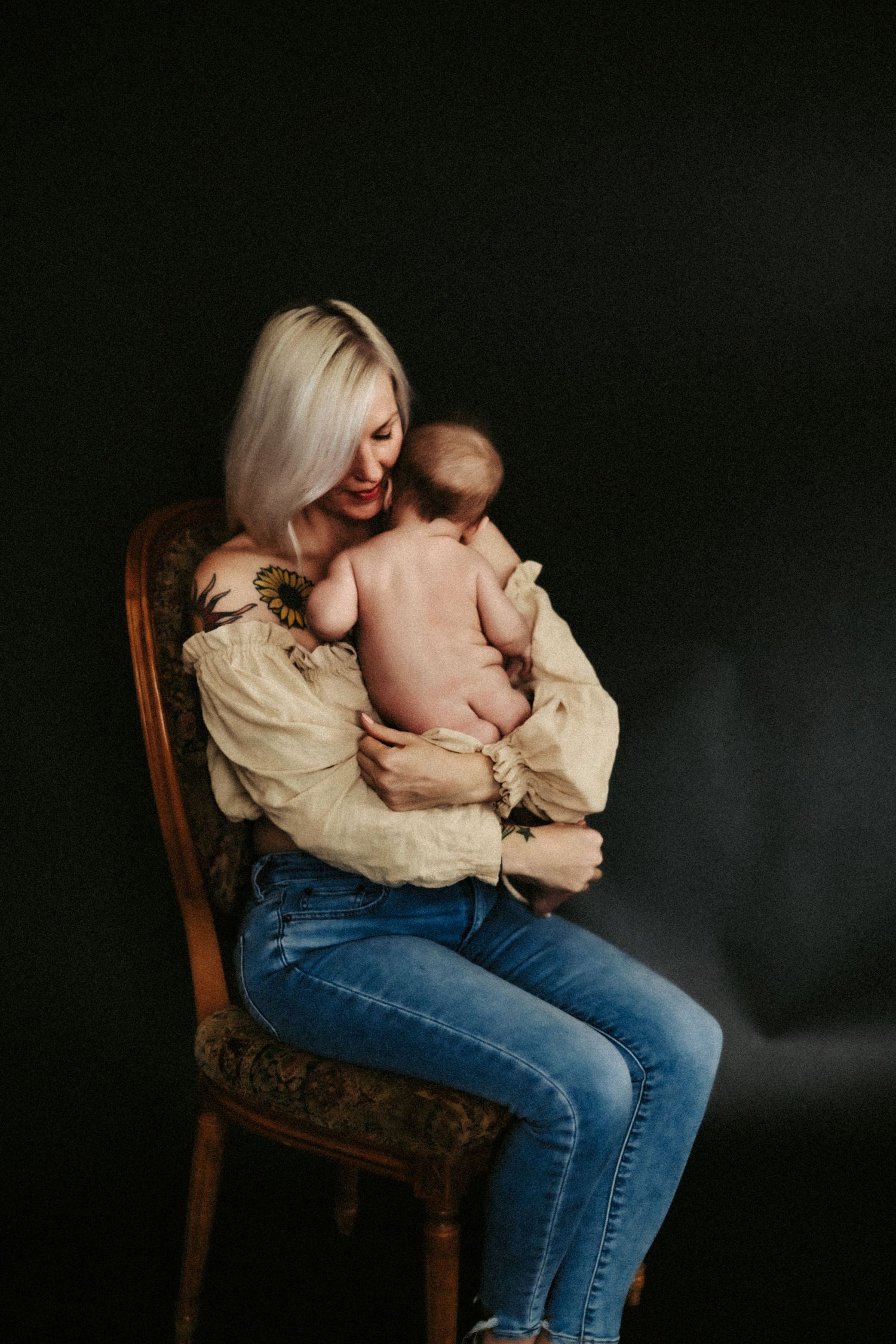 Mother in cream blouse and jeans sits on wooden chair holding bare-backed baby against dark backdrop.