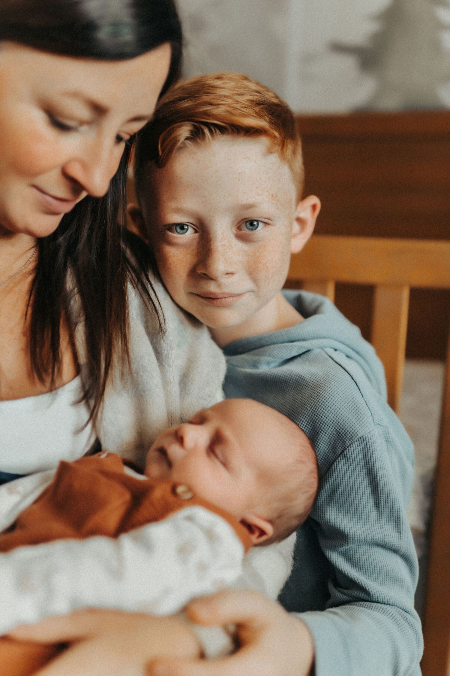 An intimate family portrait shows a mother holding a sleeping newborn while a young boy in a blue hoodie looks on.