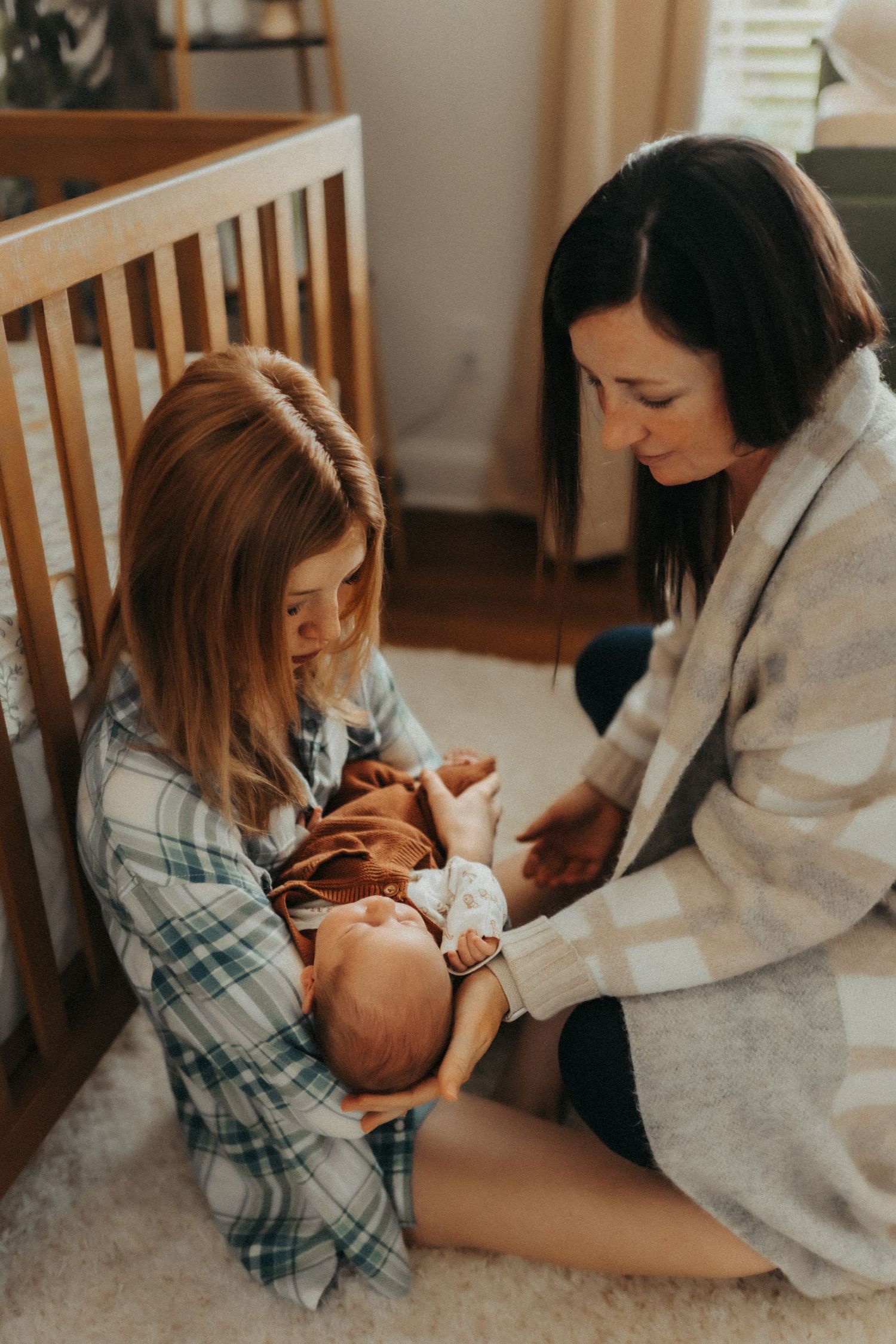 Two women sit on floor beside crib caring for a newborn baby in a cozy nursery with warm lighting.