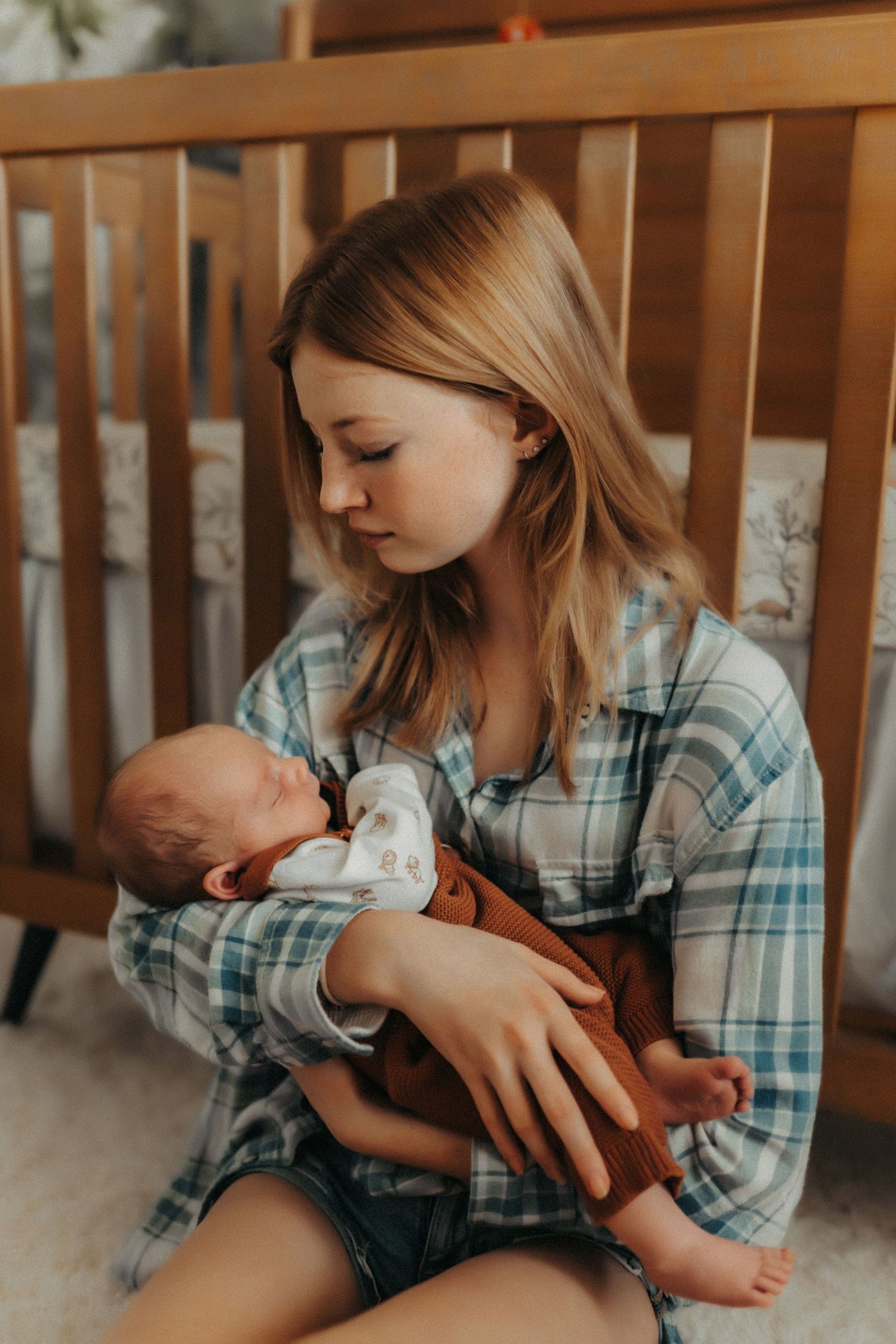 A young person in a plaid shirt tenderly holds and gazes at a newborn baby near a wooden crib.