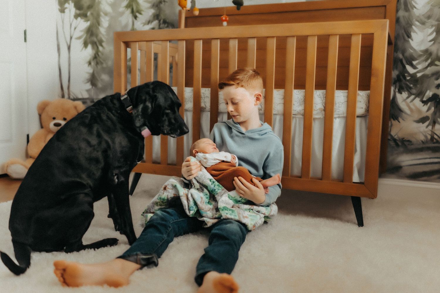 A young child sitting on the floor reading a book with a large black dog next to them by a wooden crib.