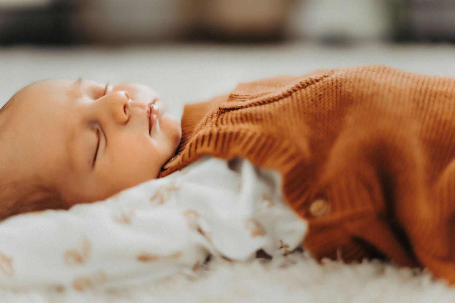Baby in a brown sweater sleeping peacefully next to a white cat on a soft surface.