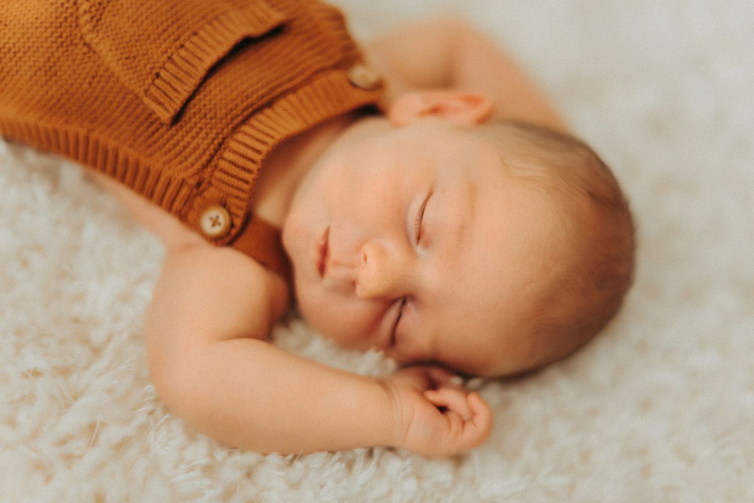 A sleeping baby wearing a brown knit sweater rests peacefully on soft white carpet.