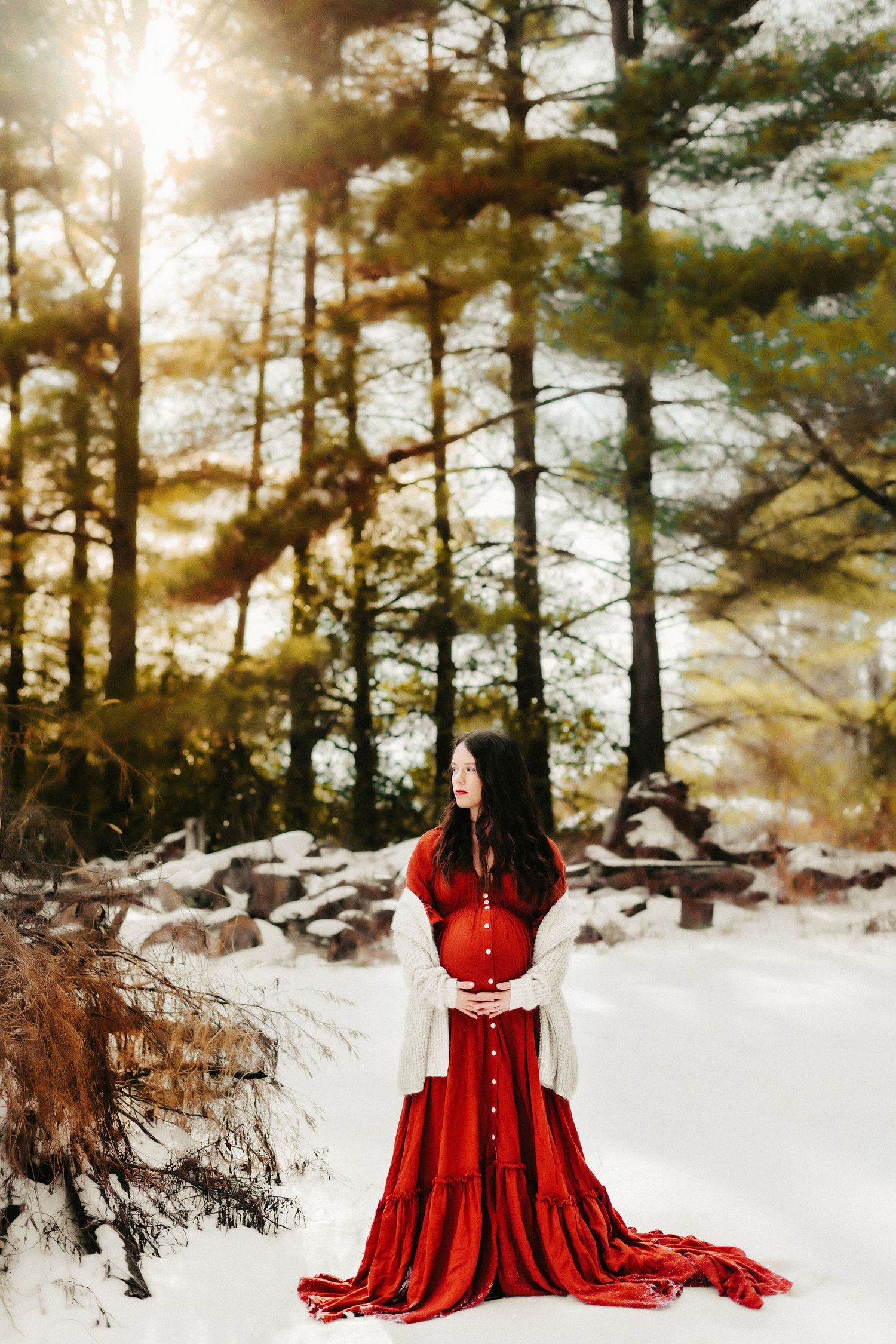 A figure in a flowing red dress stands in a snowy forest with sunlight streaming through winter pine trees.