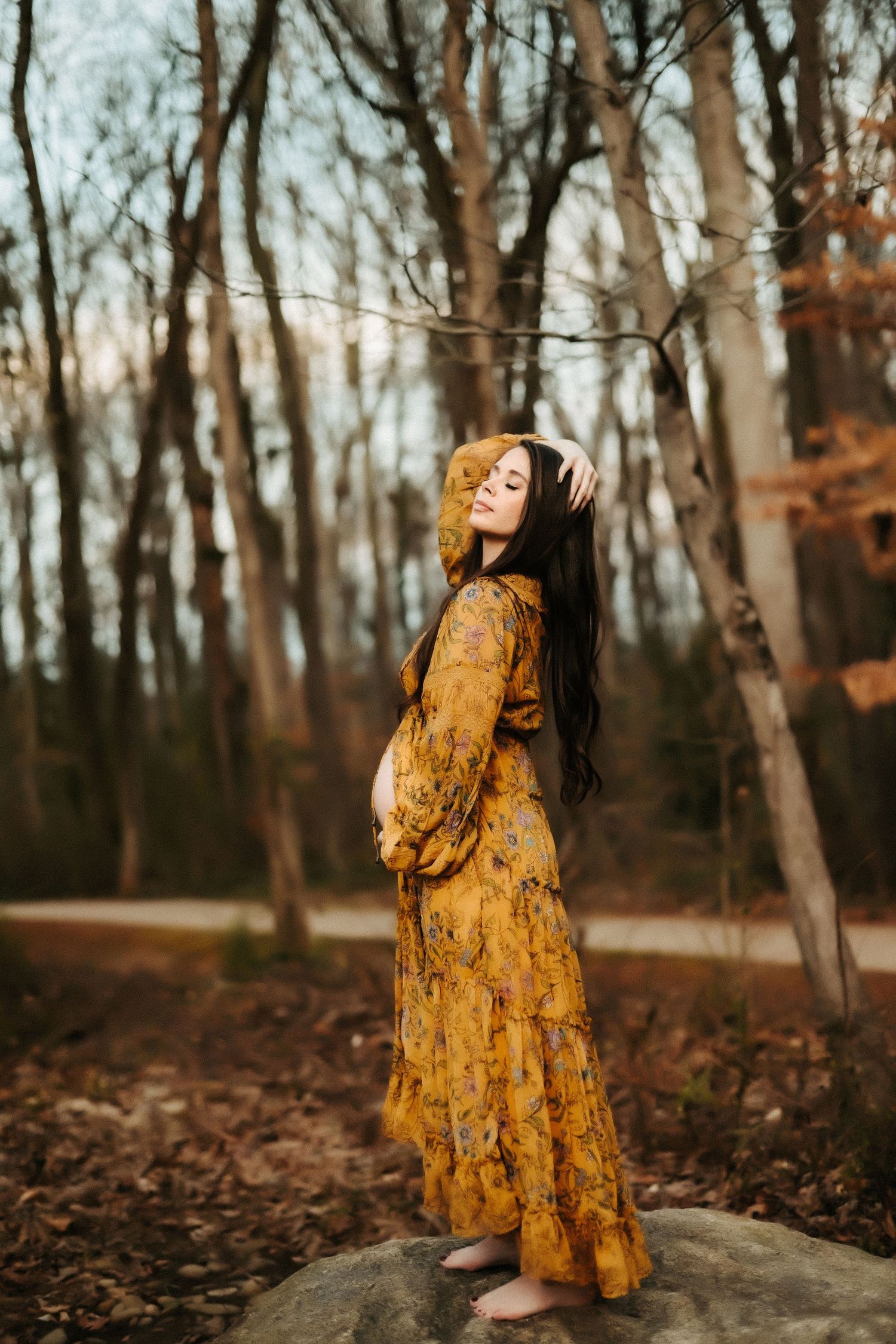 A woman in a flowing mustard yellow dress stands on a rock in an autumn forest during golden hour.