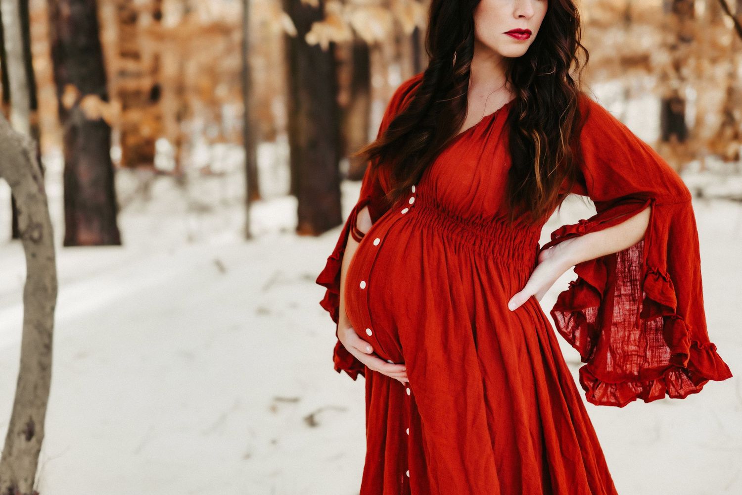 A pregnant woman in a flowing red dress poses in a snowy winter forest setting with dramatic bell sleeves.