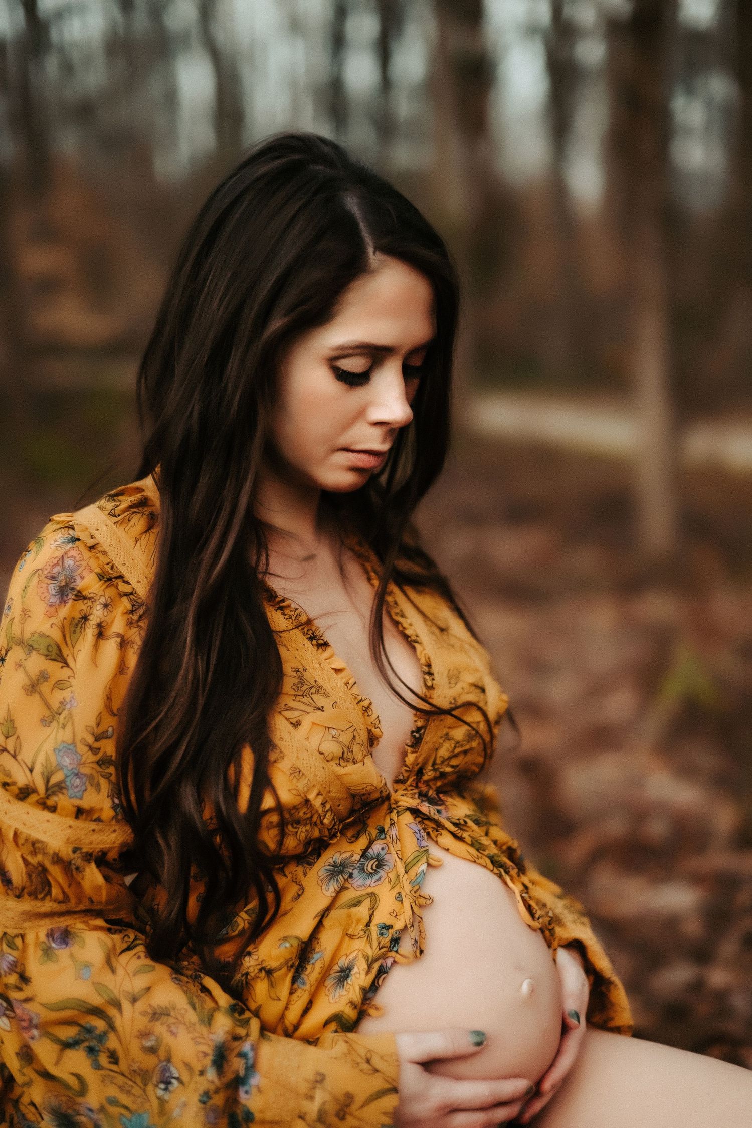 A pregnant woman in a yellow floral dress tenderly holds her baby bump while sitting in an autumn forest setting.