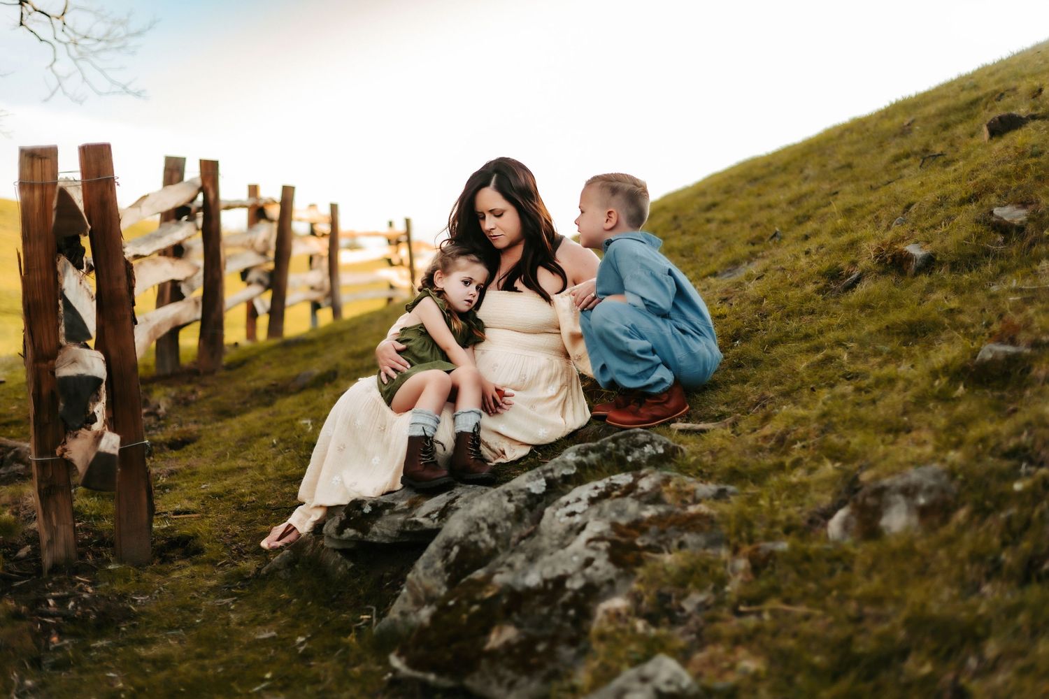 Two people sit on a large rock on a grassy hillside at sunset near a wooden fence.