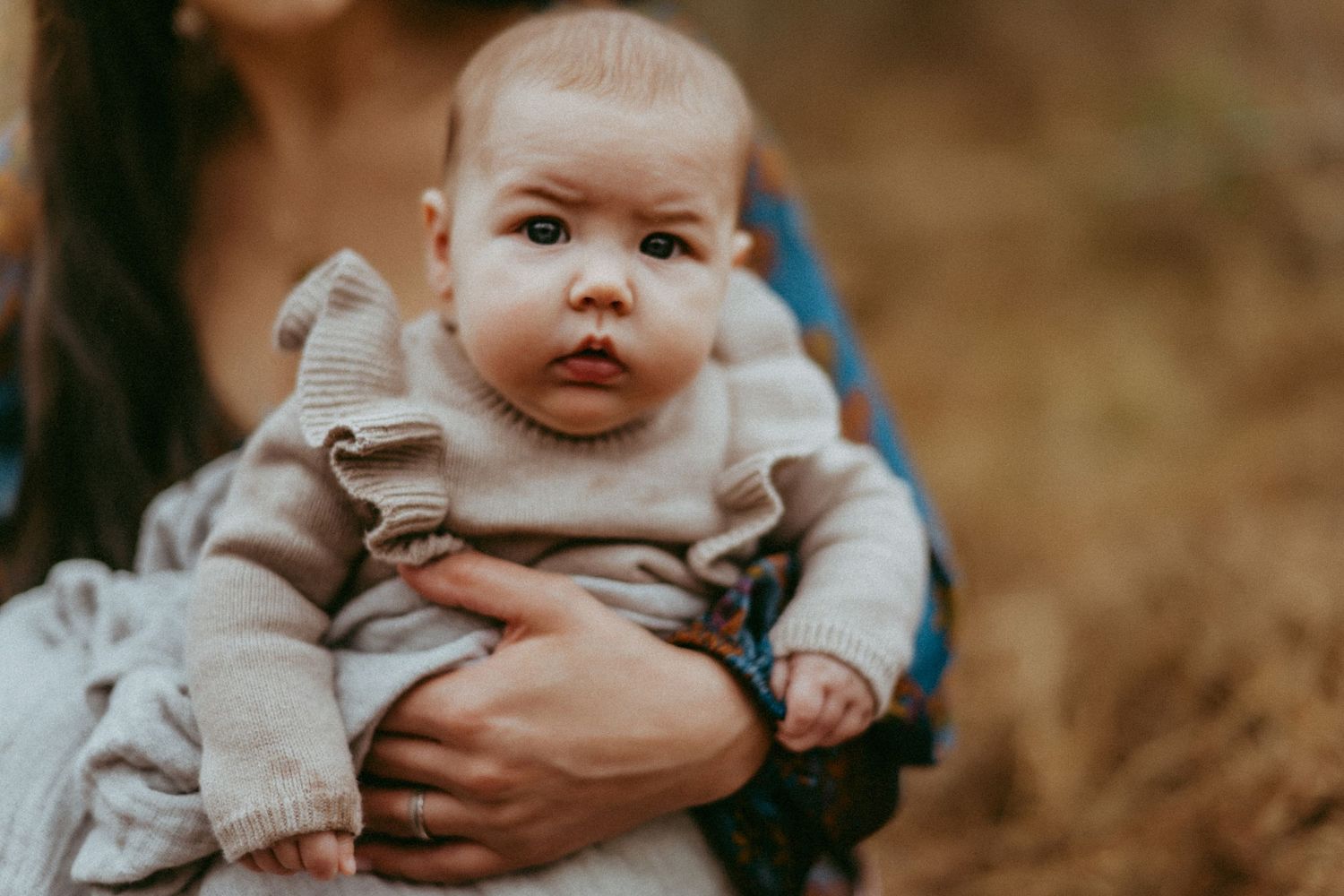 A small baby wearing a beige sweater is being held by someone against a blurred autumn background.