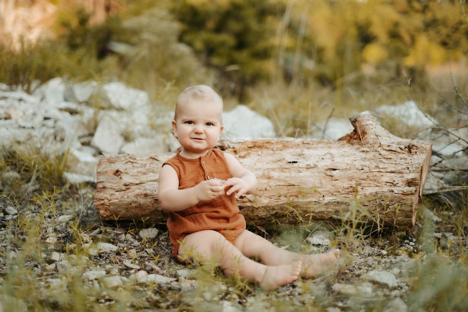 Small child in orange shirt sits on fallen log in natural outdoor setting during golden hour.