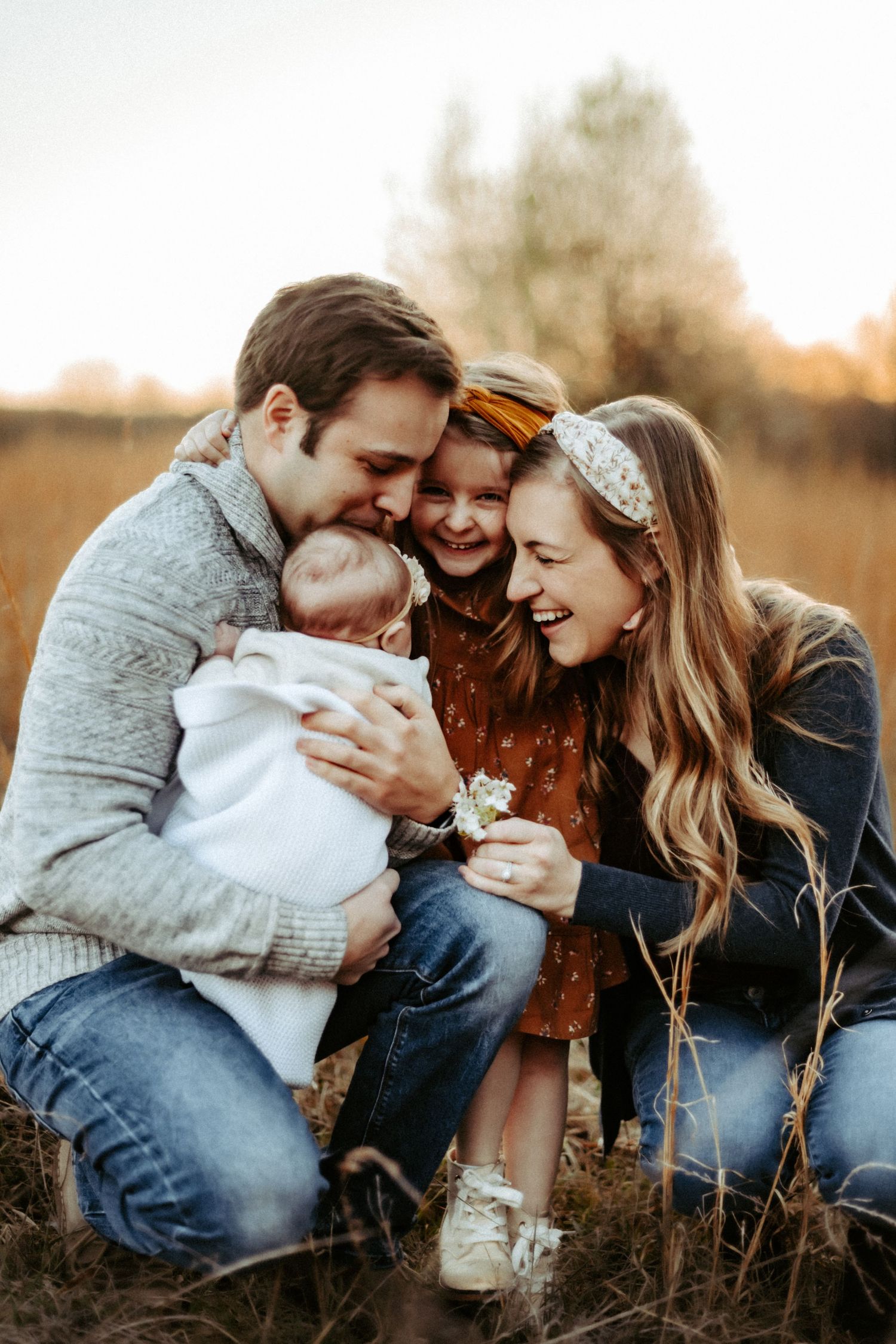 Family embraces during golden hour photo session in tall grass field.
